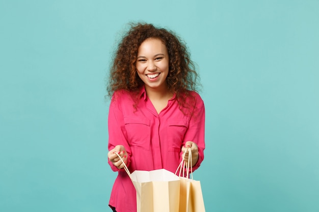 Riendo a niña africana en ropa casual rosa mantenga la bolsa del paquete con compras después de ir de compras aisladas sobre fondo de pared azul turquesa. Concepto de estilo de vida de emociones sinceras de personas. Simulacros de espacio de copia.