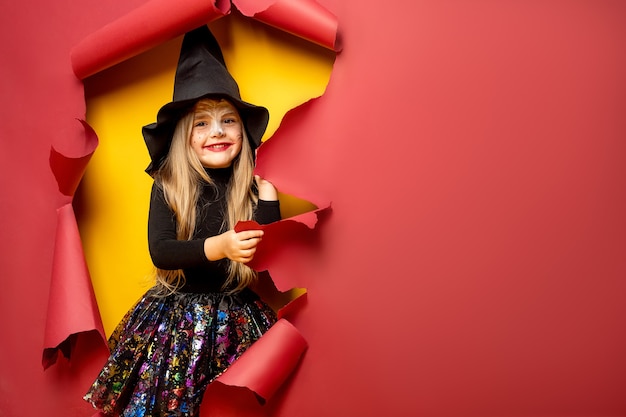 Riendo niña divertida en un disfraz de bruja de halloween mirando, sonriendo y asusta a través de un agujero de fondo de papel rojo, amarillo.