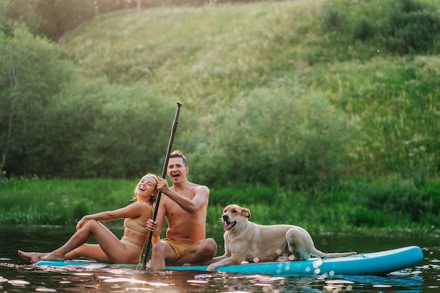 Riendo pareja caucásica sentada en paddle board con perro disfrutando de vacaciones en el río Hombre alegre