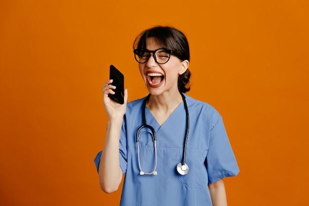 Riendo sosteniendo teléfono joven doctora vistiendo uniforme fith estetoscopio aislado sobre fondo naranja