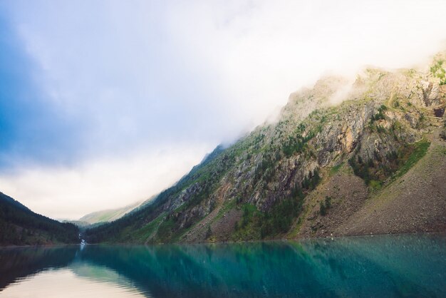 Riesige Felsen mit Bäumen im Morgennebel reflektierten sich im Wassergebirgssee. Die frühe Sonne scheint durch den Nebel. Bedecktes Wetter. Stimmungsvolle Berglandschaft majestätischer Natur.