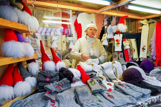 Riga, Lettland - 25. Dezember 2015: An einem der Stände während des Weihnachtsmarktes bietet und verkauft eine schöne Dame warme Kleidung, handgefertigte Souvenirs, Accessoires, Engel und Gnomenstatuen.