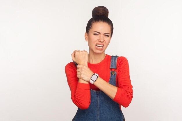 Rigidez muscular do pulso torcido Retrato de menina insalubre com coque de cabelo em macacão jeans massageando a mão dolorosa que sofre de síndrome do túnel do carpo tiro de estúdio interno isolado no fundo branco