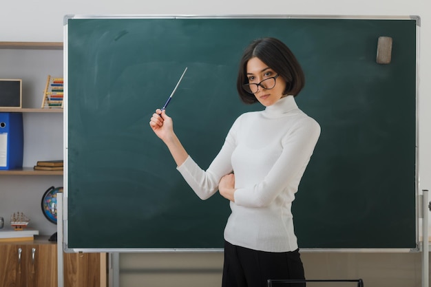 rigorosa jovem professora de óculos em pé na frente do quadro-negro e aponta para o quadro-negro com ponteiro em sala de aula