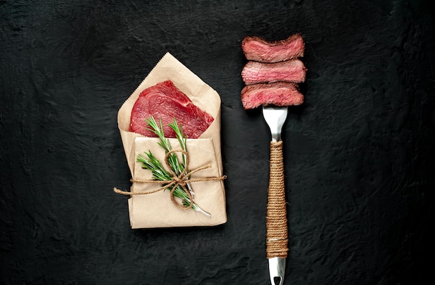 Rindersteak auf Gabel und in der Nähe von Steak in Papier auf dunklem Beton verpackt