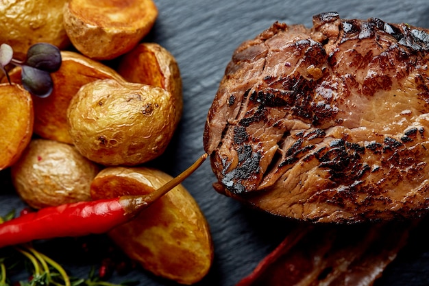 Rindersteak serviert mit Ofenkartoffeln und Gemüse, schwarze Oberfläche, Nahaufnahme. Konzept Essen.