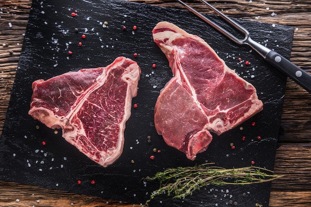 Rindersteak T-Bone. Rohes Rindersteak T-Bone auf Schieferbrett mit Salzpfefferkräutern Metzger und Gabel.