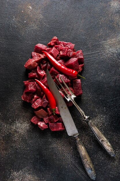 Rindfleisch mit Meersalz, getrockneten Kräutern und Chilischoten auf dunklem Schiefer- oder Betongrund in kleine Stücke schneiden. Ansicht von oben.