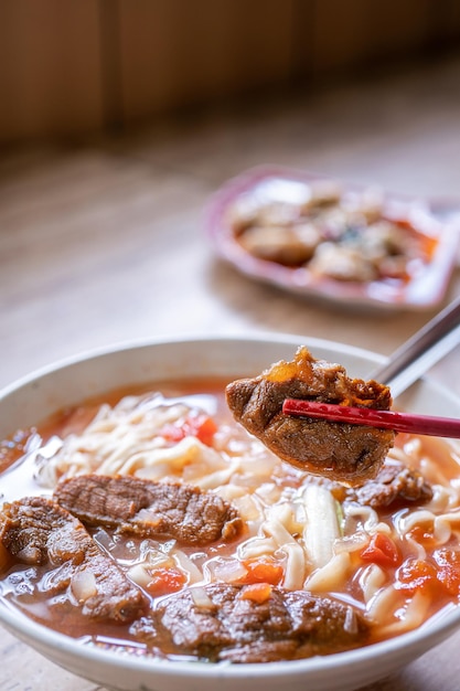 Rindfleischnudel-Taiwan-Ramen-Mahlzeit mit Tomatensauce-Brühe in Schüssel auf hellem Holztisch berühmtes Essen im chinesischen Stil, Nahaufnahme, Draufsicht, Kopienraum