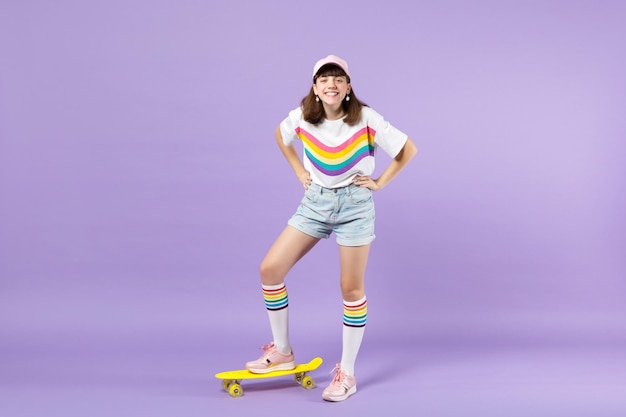 Rindo adolescente em roupas vívidas em pé com braços akimbo com skate amarelo isolado no fundo da parede pastel violeta no estúdio. Conceito de estilo de vida de emoções sinceras de pessoas. Mock up espaço de cópia.