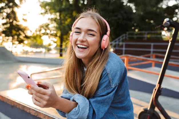 rindo alegre sorridente jovem adolescente lá fora no parque ouvindo música com fones de ouvido, segurando o telefone móvel, mostrando a língua.