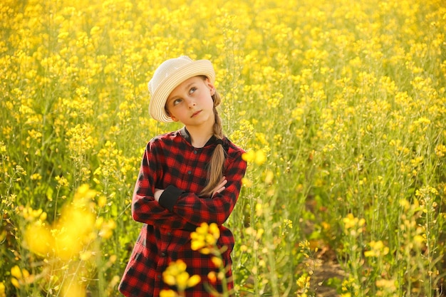 Foto rindo criança menina com chapéu e camisa em quadrados e mãos cruzadas florescendo colza amarela