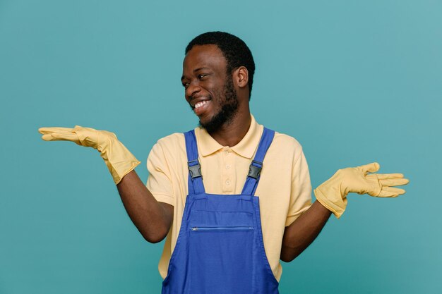 Rindo, espalhando as mãos, jovem limpador afro-americano de uniforme com luvas isoladas em fundo azul