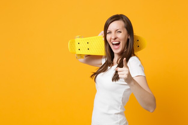 Foto rindo jovem em roupas casuais brancas, apontando o dedo indicador na câmera, segurando o skate amarelo isolado no fundo da parede laranja amarelo brilhante. conceito de estilo de vida de pessoas. mock up espaço de cópia.