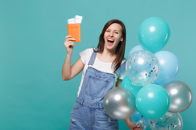 Rindo jovem em roupas jeans, segurando o bilhete de embarque passaporte comemorando com balões de ar coloridos isolados no fundo azul turquesa. Conceito de emoções de pessoas de festa de aniversário.