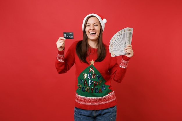 Rindo jovem garota de Santa segurando o cartão de crédito, muitos monte de notas de dólares, dinheiro isolado em fundo vermelho brilhante. Conceito de festa de feriado de celebração de ano novo feliz 2019. Simule o espaço da cópia.