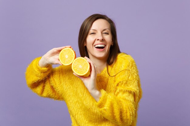 Rindo jovem na camisola de peles segurando nas mãos metades de frutas frescas laranja maduras isoladas em fundo pastel violeta no estúdio. O estilo de vida vívido das pessoas relaxa o conceito de férias. Mock up espaço de cópia.