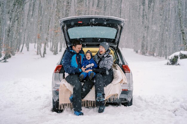 Rindo mãe pai e menina estão sentados em um cobertor na bagageira de um carro sob uma queda de neve em