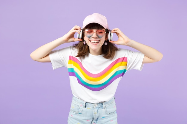 Rindo menina adolescente em roupas vívidas, óculos ouvem música com fones de ouvido, mantendo os olhos fechados isolados em fundo pastel violeta. Emoções sinceras de pessoas, conceito de estilo de vida. Mock up espaço de cópia.