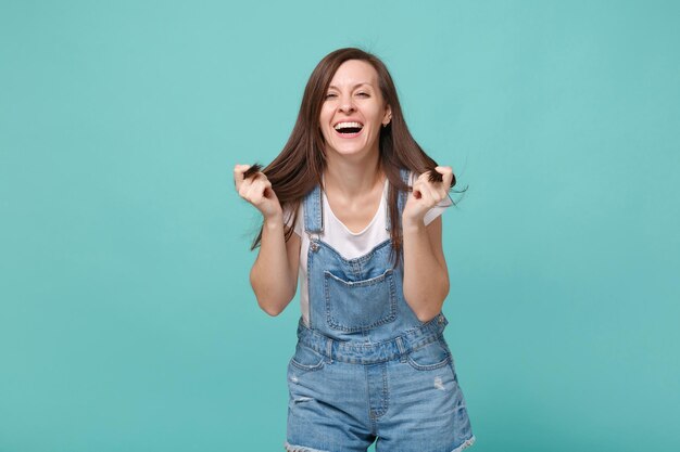 Rindo menina jovem morena em roupas jeans casuais posando isolado no retrato de estúdio de fundo azul turquesa. Conceito de estilo de vida de emoções sinceras de pessoas. Simule o espaço da cópia. Segurando o cabelo.