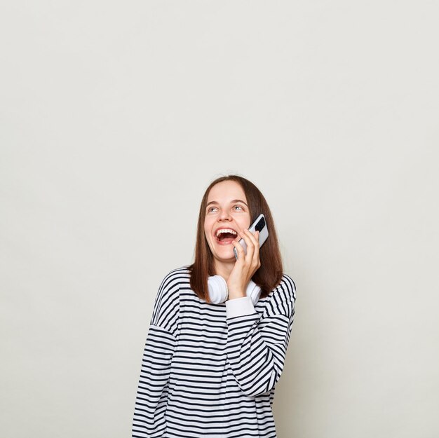 Rindo mulher de cabelos castanhos vestindo camisa listrada posando isolada sobre fundo cinza falando no telefone celular copie o espaço antes da maquete de anúncio