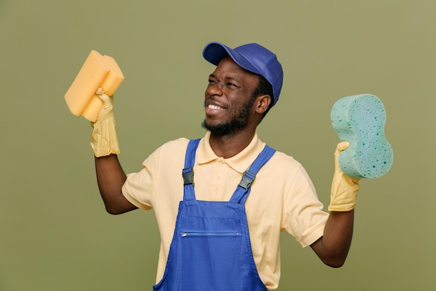 Rindo segurando esponjas de limpeza jovem limpador afro-americano de uniforme com luvas isoladas em fundo verde