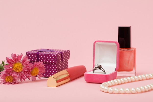 Ring in einer Box, Nagellack, Lippenstift, Perlen, Geschenkbox und Blumen auf rosa Hintergrund. Damenschmuck, Kosmetik und Accessoires.