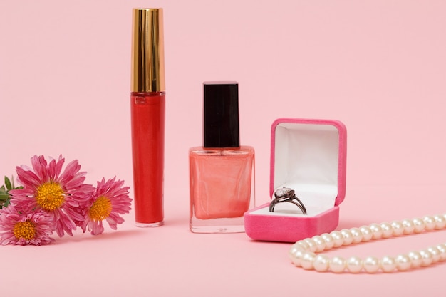 Ring in einer Box, Nagellack, Lippenstift, Perlen und Blumen auf rosa Hintergrund. Damenschmuck, Kosmetik und Accessoires.