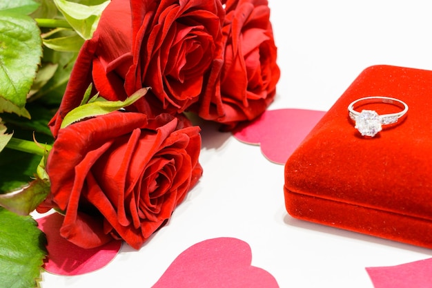 Ring mit Diamant auf Geschenkbox und Strauß roter Rosen und Papierherzen auf weißem Hintergrund