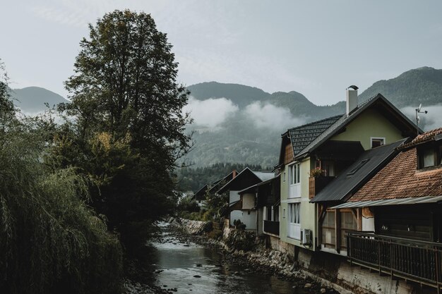 Rio de montanha e fileira de casas perto dele na cidade eslovena Bohinjska Bistrica sob os Alpes Julianos escondidos nas nuvens ao fundo durante o nascer do sol durante a manhã de verão