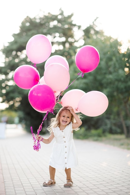 Foto rir engraçado criança menina de 3-4 anos segurando balões cor-de-rosa usar vestido branco ao ar livre sobre a natureza