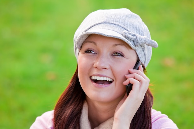 Risa mujer joven con gorra y bufanda hablando por teléfono en la hierba