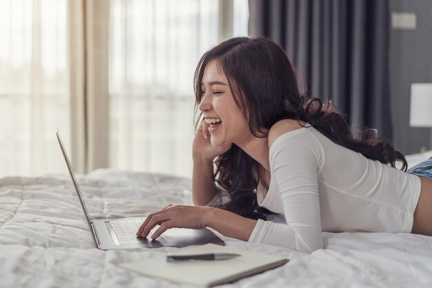 Risa mujer joven tumbado en la cama y usando la computadora portátil