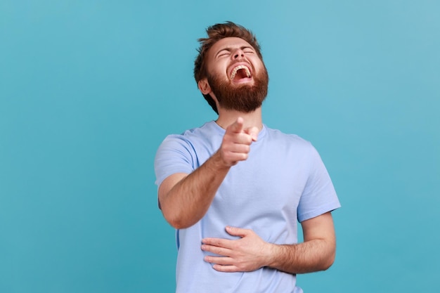 Risos hilariantes retrato de homem barbudo feliz alegre rindo alto e apontando para a câmera zombando provocando você segurando a barriga foto de estúdio interior isolada em fundo azul