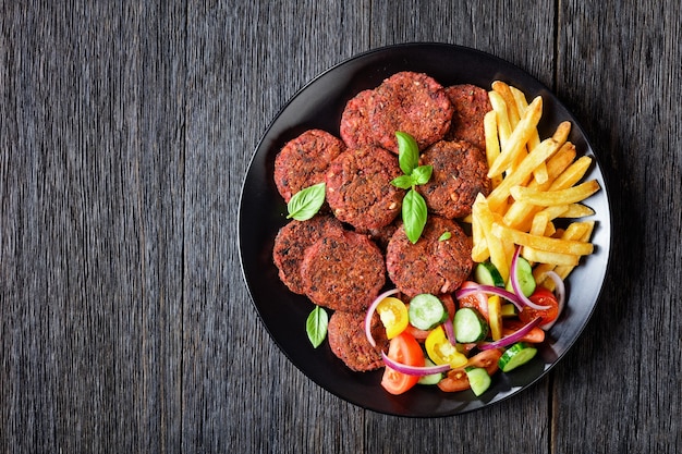 Rissóis de beterraba com cogumelos e feijão preto servidos com batatas fritas e uma salada de vegetais de tomate, pepino, cebola roxa e manjericão fresco em um prato preto sobre uma mesa de madeira escura, vista de cima, espaço livre