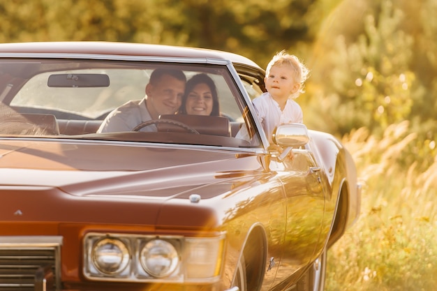 Riviera en estilo retro en puesta de sol. coche único lindo niño rubio está sentado al volante de un coche retro con su familia. los padres están sentados en el asiento trasero, el niño se asoma por la ventana.