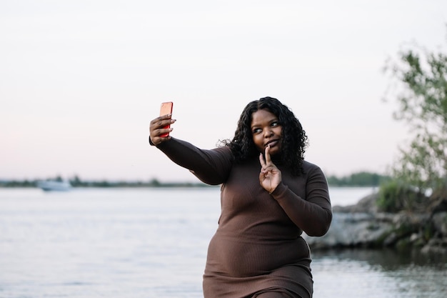 Rizado riéndose bastante mujer afroamericana en vestido elegante tomar selfie por teléfono inteligente al lado del mar Copiar espacio