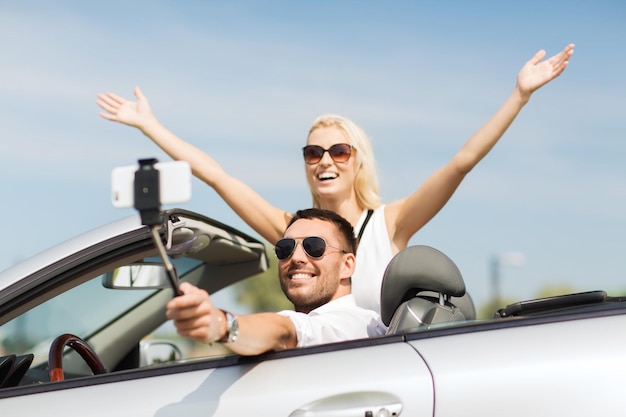 road trip, freizeit, paar, technologie und personenkonzept - glücklicher mann und frau, die im cabriolet fahren und mit dem smartphone auf dem selfie-stick fotografieren