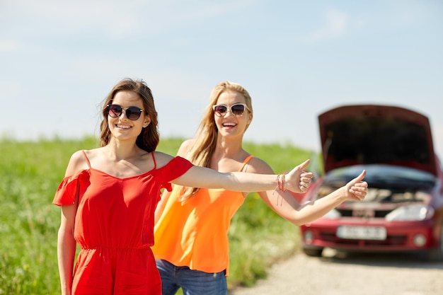 Road Trip, Transport, Reisen, Gesten und Personenkonzept - glückliche junge Frauen mit kaputtem Auto, die per Anhalter Geste zeigen und auf dem Land um Hilfe bitten