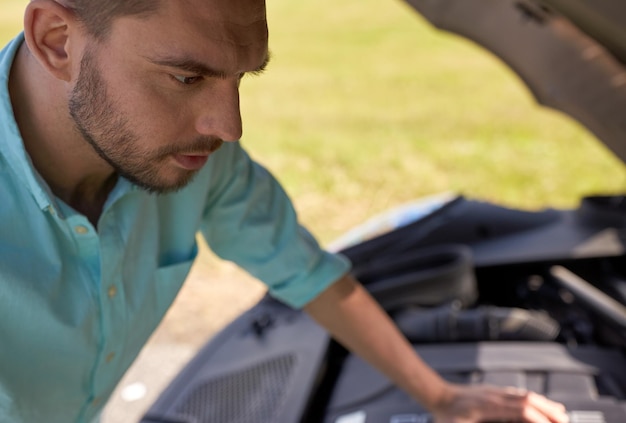 Foto roadtrip-, transport-, reise- und personenkonzept - nahaufnahme eines jungen mannes mit offener motorhaube eines kaputten autos auf dem land