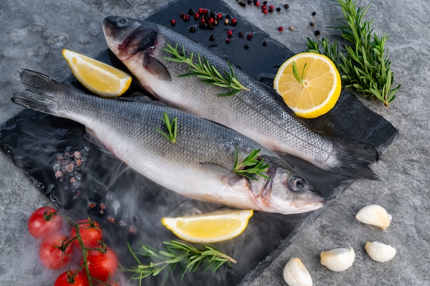 Robalo cru fresco com tomate limão e especiarias na placa de pedra preta com névoa de vapor frio de geada. Frutos do mar de alimentos frescos no conceito de mercado.