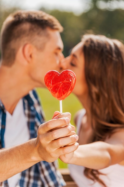 Robar un beso detrás de la piruleta. Pareja amorosa escondida detrás de piruleta en forma de corazón rojo mientras ambos de pie al aire libre