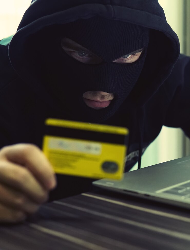 Foto robo de internet: un hombre que usa un pasamontañas y sostiene una tarjeta de crédito mientras está sentado detrás de una computadora portátil, fondo blanco.