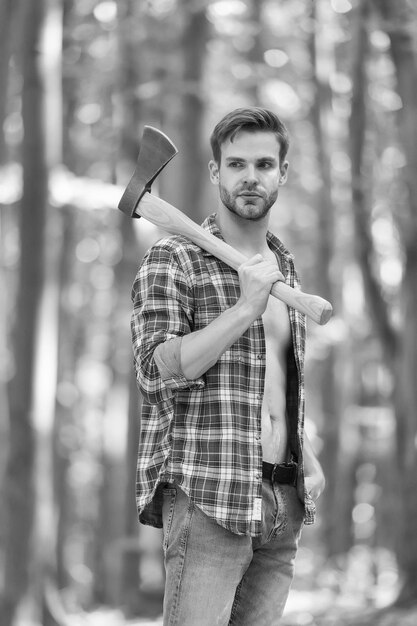 Robuster Look Unrasierter Mann trägt Axt im Wald Holzfäller-Stil Axtmann trägt offenes kariertes Hemd mit Jeans Lässiger Stil Naturreisen Sommermode Trendiger Stil Alltagsgarderobe