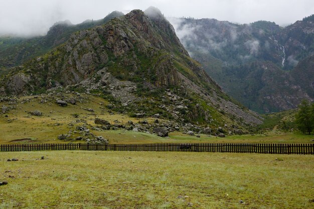 Roca envuelta en niebla, cubierta de pasto verde, valla de madera debajo de la montaña