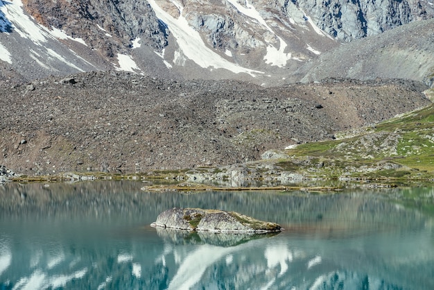 Roca en lago de montaña turquesa. Montaña nevada reflejada en el agua clara azul del lago glacial. Hermoso fondo soleado con reflejo de glaciar blanco como la nieve en la superficie del agua verde del lago de montaña.