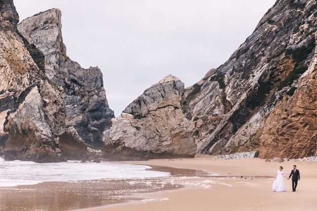 Rocas altas cerca del océano y clima nublado. los recién casados se dan la mano y caminan junto al mar.