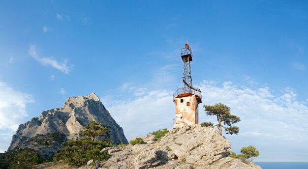 Rocha com árvores coníferas e antigo farol na encosta no fundo do céu azul (rocha "Sokol", reserva "Novyj Svit", Crimeia, Ucrânia). Imagem de costura de cinco tiros.