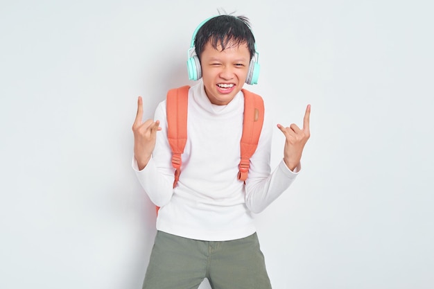 Rock and Roll guapo joven estudiante asiático con sombrero de Navidad y ropa informal con mochila escuchando música rock con auriculares y mostrando gesto de rock metálico aislado en fondo blanco