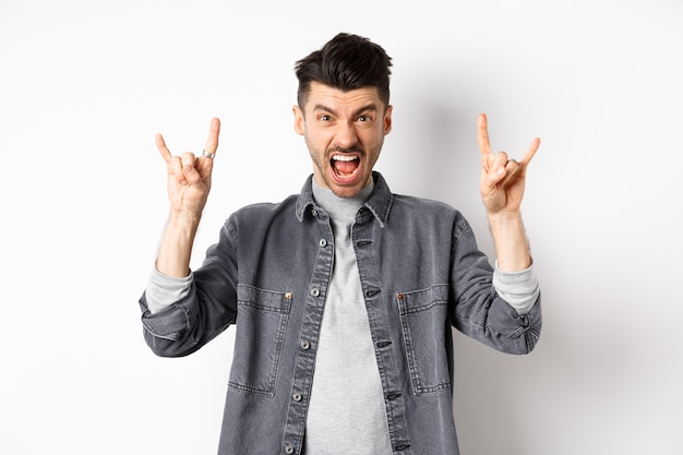 Rock on. Joven descarado gritando y mostrando el signo de cuernos de heavy metal, asistir a un concierto impresionante, divertirse, de pie sobre fondo blanco.